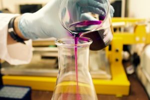 photo of a chemistry lab with a close up of a white gloved hand pouring a dark pink liquid into the top of a glass flask.