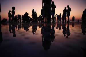 Photograph of people of different ages standing in shallow water. The sun is setting over the water.